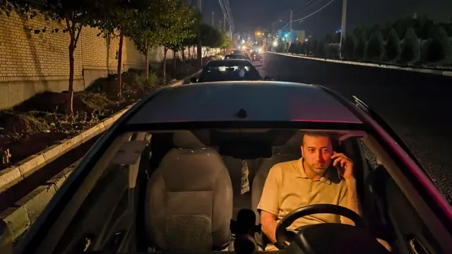 A man sits in his car on the phone, a long line of cars behind him