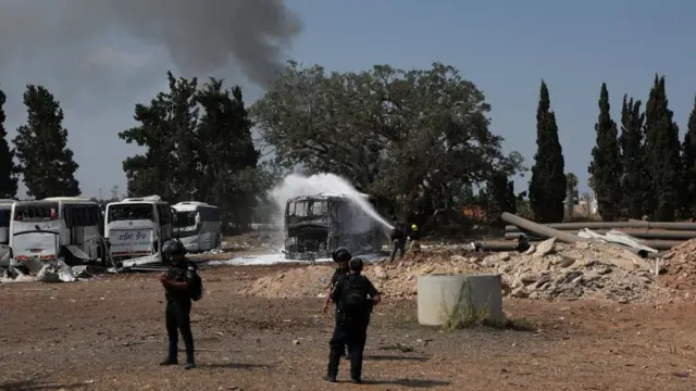 Firefighters spray water on a burnt-out bus with smoke on the horizon