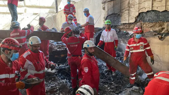 Around a dozen Red Crescent rescuers standing in the rubble doing various things. They are wearing red and white uniforms and hard hats. Some are wearing surgical face masks and other orange work gloves
