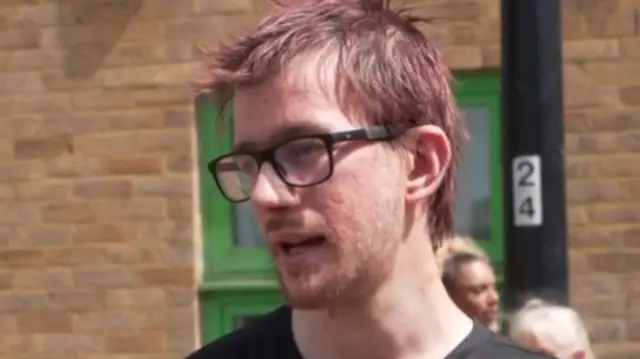 Bradley Latter while speaking with reporters. He has short red hair and wears glasses and a black T-shirt.