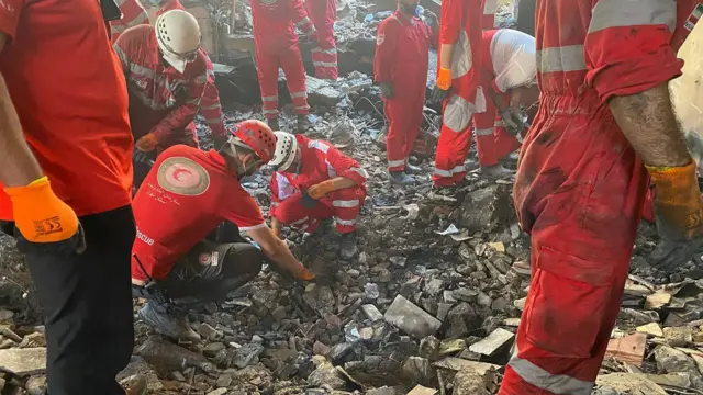 Multiple Red Crescent staff working in the rubble. There are two crouched down.