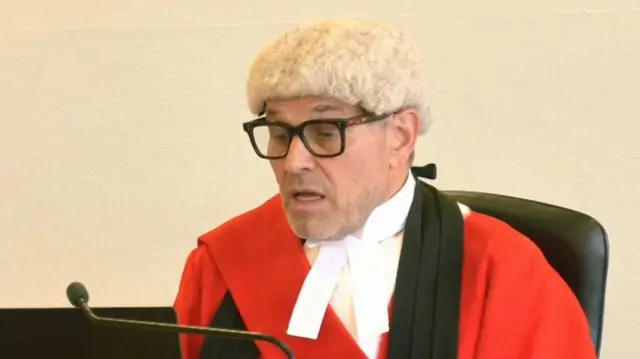 Judge Kerr in red, white and black judge's robes. He has a white wig and is sitting at his desk in the courtroom.