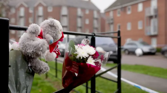 A floral tribute is tied to a fence on a residential street. A teddy has also been tied next to it.
