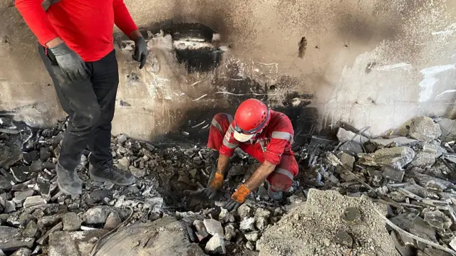 Red Crescent member wearing red uniform, red hard hat and orange work gloves crouching and in rubble and investigating, while anothert is standing on the left and pointing. the wall behind him is scorched