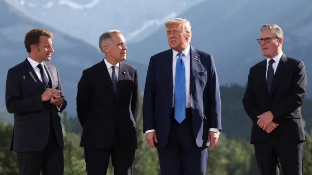 Emmanuel Macron, Mark Carney, Donald Trump and Keir Starmer speak. There are mountains behind them and a forest is seen. They are outside at the G7 summit for the annual family photo.