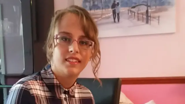Teohna Grant smiles as she sits in a chair. She has light brown hair that is tied back with some hair framing her face. She wears glasses and a chequered shirt.