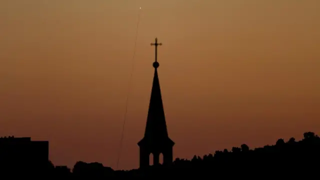 From a distance, a missile is seen rising into an orange sky over the steeple of a church