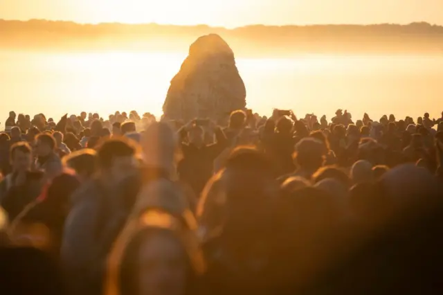A large crowd of people are gathered for sunrise at Stonehenge. The foreground is a sea of blurred heads which are shadowed, with one of the monument's large stones in the background. The sky is a light orange hue.