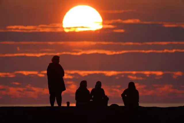 Three people are sat down watching the sun rise and one person is standing up. The sun is rising between clouds in front of them.