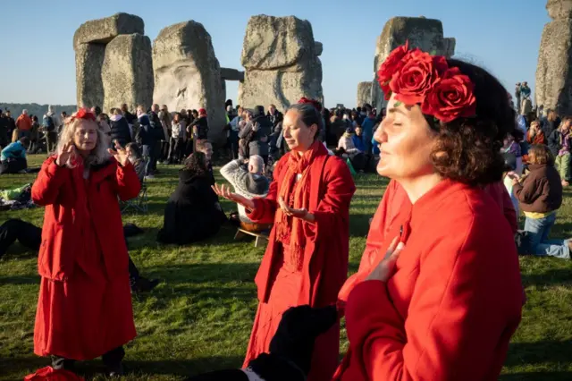 Three revellers at Stonehenge during the 2024 Summer Solstice. The three women are standing together and are wearing long red dresses and red flowers in their hair. They all have their eyes closed, one has her hand clasped to her chest while the others have their palms out. Their faces are bathed in sunlight and the ancient stones and a crowd can be seen behind them.