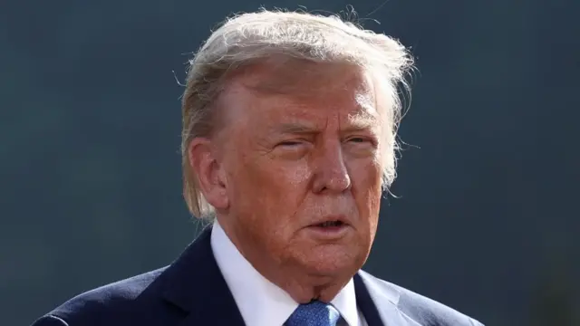 donald trump looks straight ahead with his mouth slightly open. he has grey-blond hair and is wearing a blue suit. it is a head and shoulders photo.