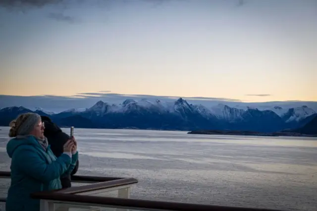 Woman in a thick turquoise coat holds her phone up to capture the midnight sun among the snowy Norwegian mountains.