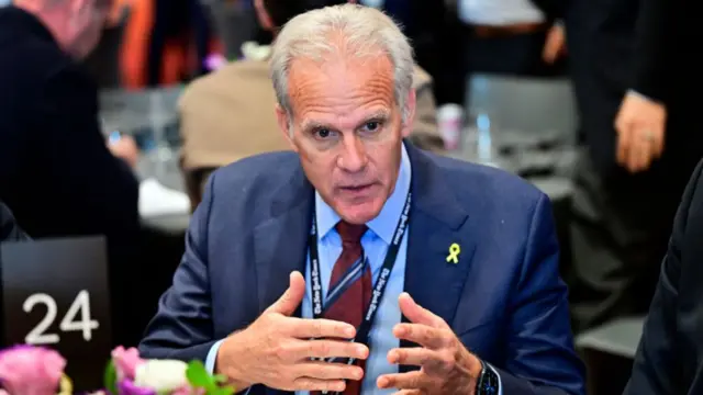 A white man in a suit gestures as he speaks.