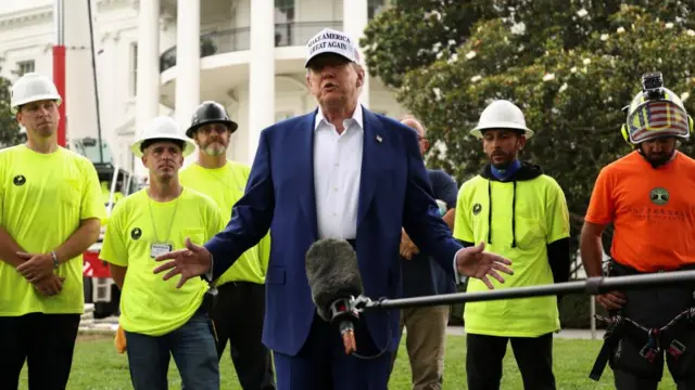 Donald trump, flanked by workers in high vis, speaks to press on the white house lawn.