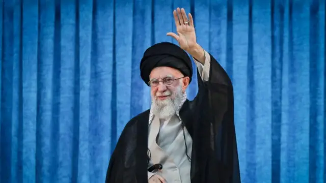 An elderly bearded man lifts a hand to wave in front of a blue curtain.