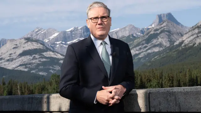 Starmer wears a dark suit and stands against a backdrop of mountains and forestry