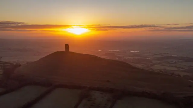Glastonbury Tor - a hill with a tower on top. In the background, the sun rises