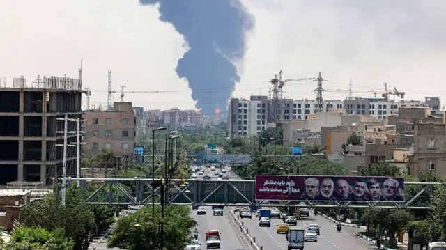 Portraits of Iranian military generals and nuclear scientists, killed in Israel's June 13 attack are displayed above a road, as a plume of heavy smoke and fire rise from an oil refinery in southern Tehran, after it was hit in an overnight Israeli strike
