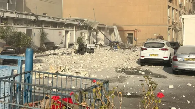 A building with debris on the ground, cars nearby.