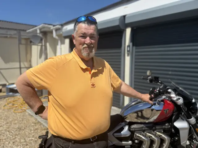 Lee Corbet in a yellow T-shirt stood by a large motorcycle