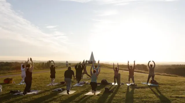 Several people partake in a yoga session in front of the Light Pyramid in Milton Keynes. They are all in a lunge position and have their hands raised above their heads towards the sky.