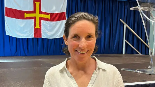 Lindsay de Sausmarez standing in front of a stage decorated with Guernsey flags