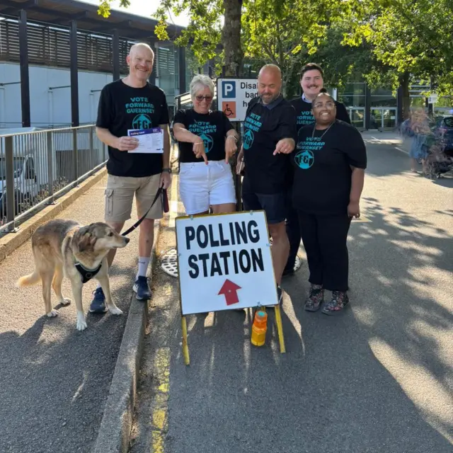 Five people stood by a polling sign with a dog