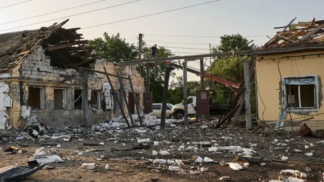 A single-floor building is significantly damaged, with its roof blown out in several places and entire walls missing. Rubble surrounds the site.