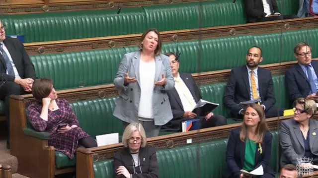Ellie Chowns in light grey blazer and matching jeans stands in House of Commons, green leather benches behind her and colleagues sitting to her right and left