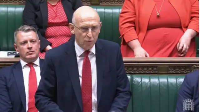 John Healey in dark suit, white shirt and red tie stands in front of House of Commons's green leather benches