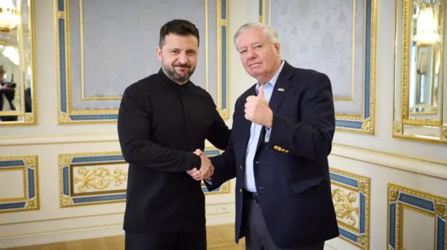 Volodymyr Zelensky (L) in black shirt and trousers as he politely smiles while shaking hands with Senator Lindsey Graham (R) in a tie-less black suit and a light blue shirt. He's holding his left arm up and giving a thumbs up. Behind them an ornate wall inside the Ukrainian presidential palace