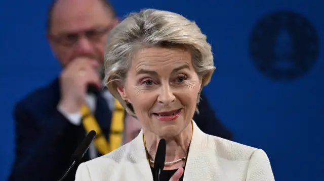 Ursula von der Leyen, a woman with short blonde hair wearing a white jacket, speaks into a small microphone in front of a blue background