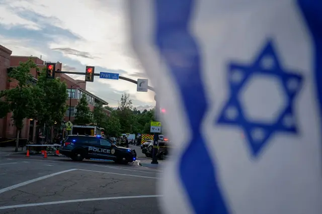 Israeli flag waves in foreground as police and police tape are seen in the background of a street