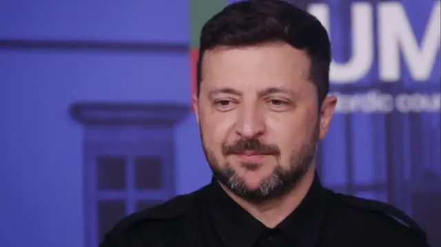 Close up of Volodymyr Zelensky in short-sleeved black top with blue wall in background