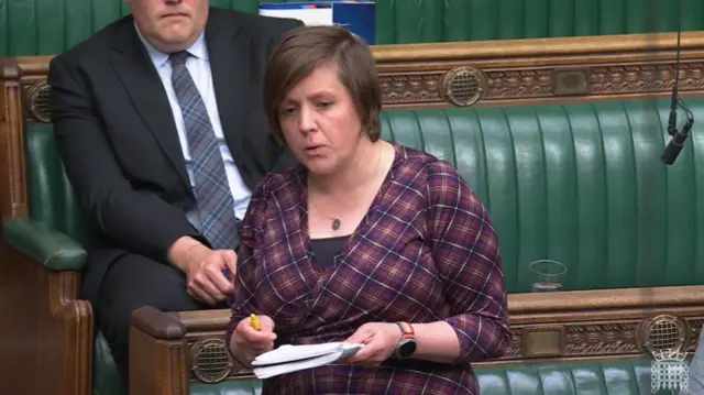 SNP MP Kirsty Blackman in burgundy tartan dress with blue and yellow chequers stands in house of Commons. She has short brown hair, a necklace around her neck and a watch around her left wrist. She's holding a pen and notebook