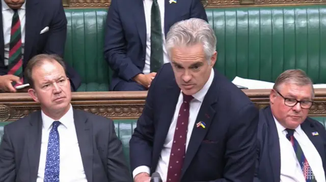 James Cartlidge stands in front of despatch box in black suit, white shirt and burgundy tie, to his left, sitting on green leather bench is MP Mark Francois
