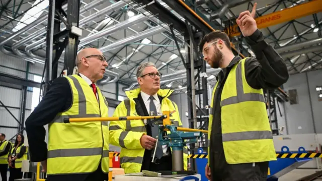 John Healey (L) and Keir Starmer (C) listen to a BAE employee (R) speaking as they visit the defence firm's headquarters in Glasgow. All three are wearing yellow hi-vi jackets while inside what looks like a hangar