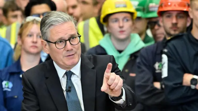 Headshot of Starmer talking while raising one hand and pointing with his index finger. People stand behind him