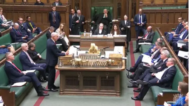Wide shot on inside of House of Commons with MPs seen sitting on green leather benches, John Healey standing as he addresses the Opposition