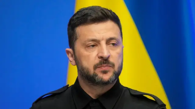 Close up of Volodymyr Zelensky in a black shirt as he speaks during press event, a blue wall background and the Ukrainian flag behind him