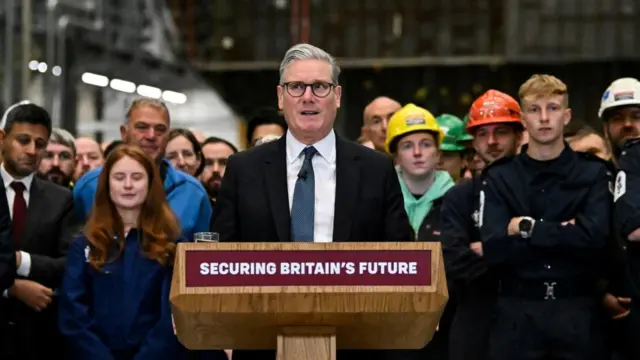 Starmer talking at a podium which says Securing Britain's Future on the front of it. Workers stand behind him