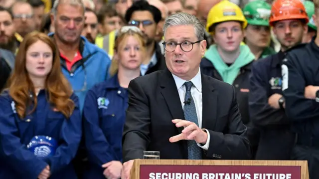 Keir Starmer in black suit, white shirt and blue tie delivers speech at lectern, a group of workers in uniform behind him