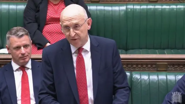 JOhn Healey stands in Parliament wearing dark suit, red tie and white shirt as he delivers speech. Behind him are Commons' green leather benches on which some of his colleagues are sitting