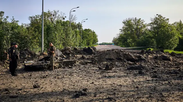 Three policemen in uniform stand on a road covered in debris and rubble from the strike.
