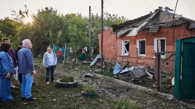 Six people stand dotted around a building with its three windows blown out and significant damage to its roof.