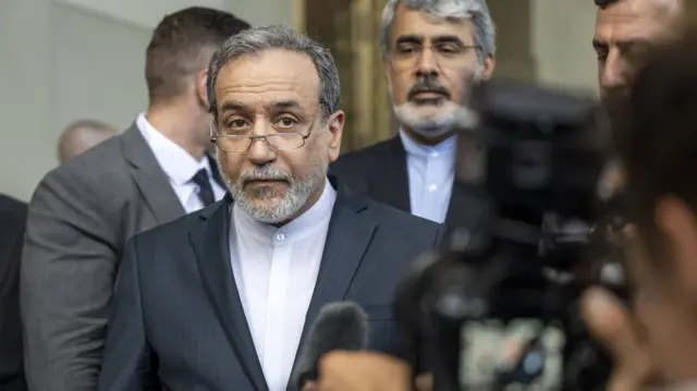 Abbas Araghchi walks towards someone from the media holding a camera facing him. He has a grey beard and black hair and brown eyes. He is wearing a white shirt, black blazer, and silver-rimmed glasses.