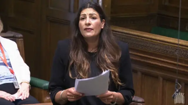 Preet Kaur Gill in black blazer and shirt, holds stack of paper as she speak, part of the Common's wooden benches visible behind her