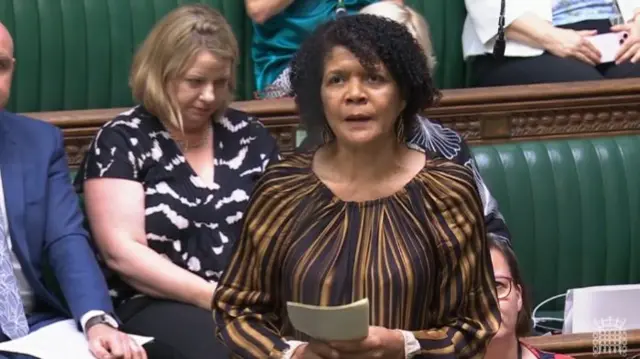 Chi Onwura stands in beige and dark brown top, colleagues on leather green benches behind her