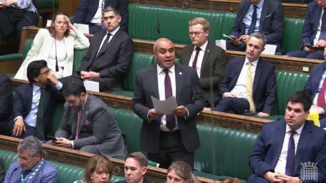 Labour MP David Burton-Sampson (centre) stands in a dark grey suit, white shirt and burgundy tie, papers clutched in his hands, several MPs sitting on the Commons' green leather benches around him