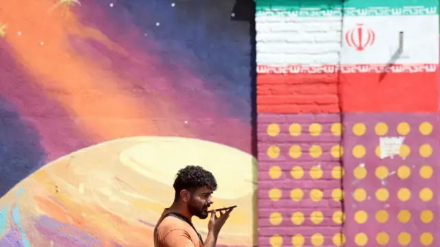 An Iranian man speaks on his phone as he walks past a mural on a wall.